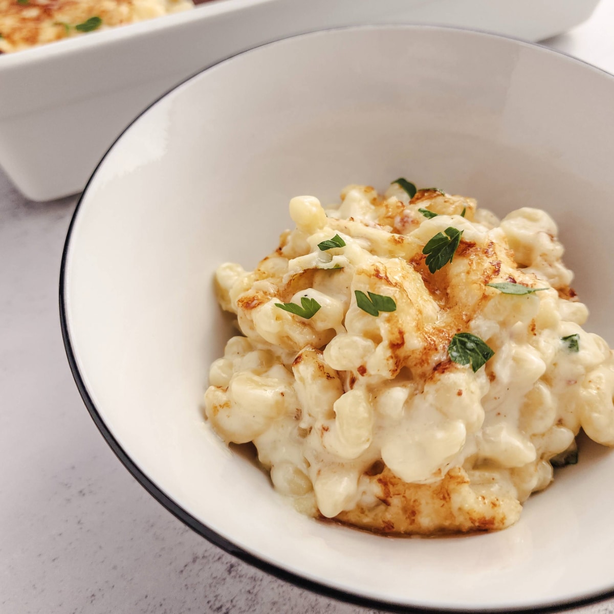 A white bowl of macaroni and cheese, garnished with fresh parsley on a stone counter.