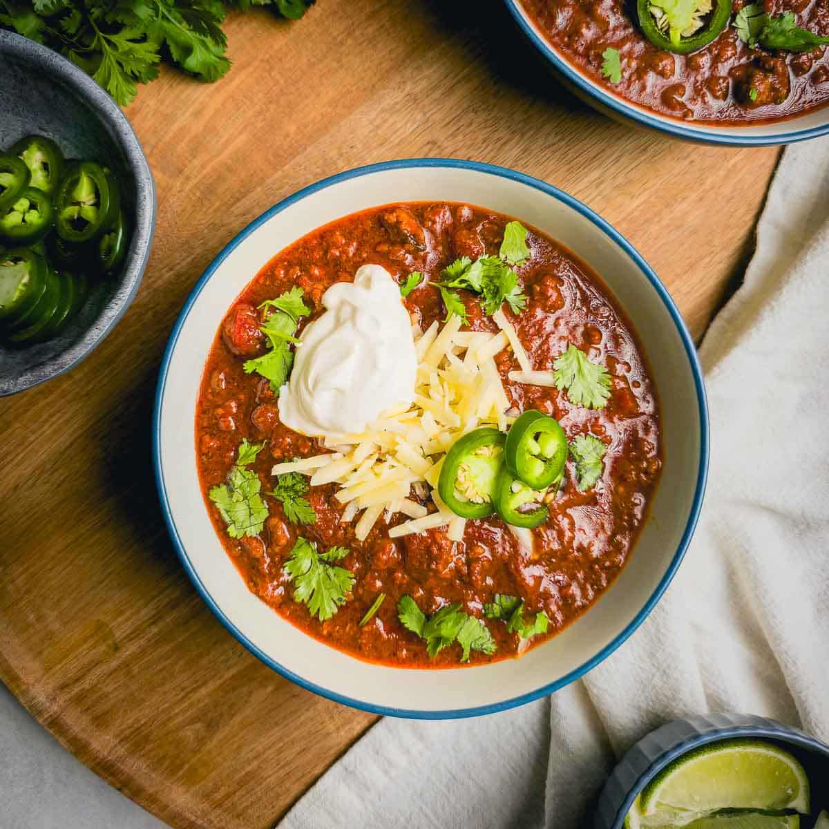 A bowl of garnished beef and chorizo chili in a white bowl on a wooden cutting board.
