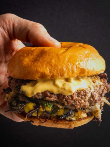 A hand holding a smash burger with onion and jalapeño, topped with pickled okra burger sauce.