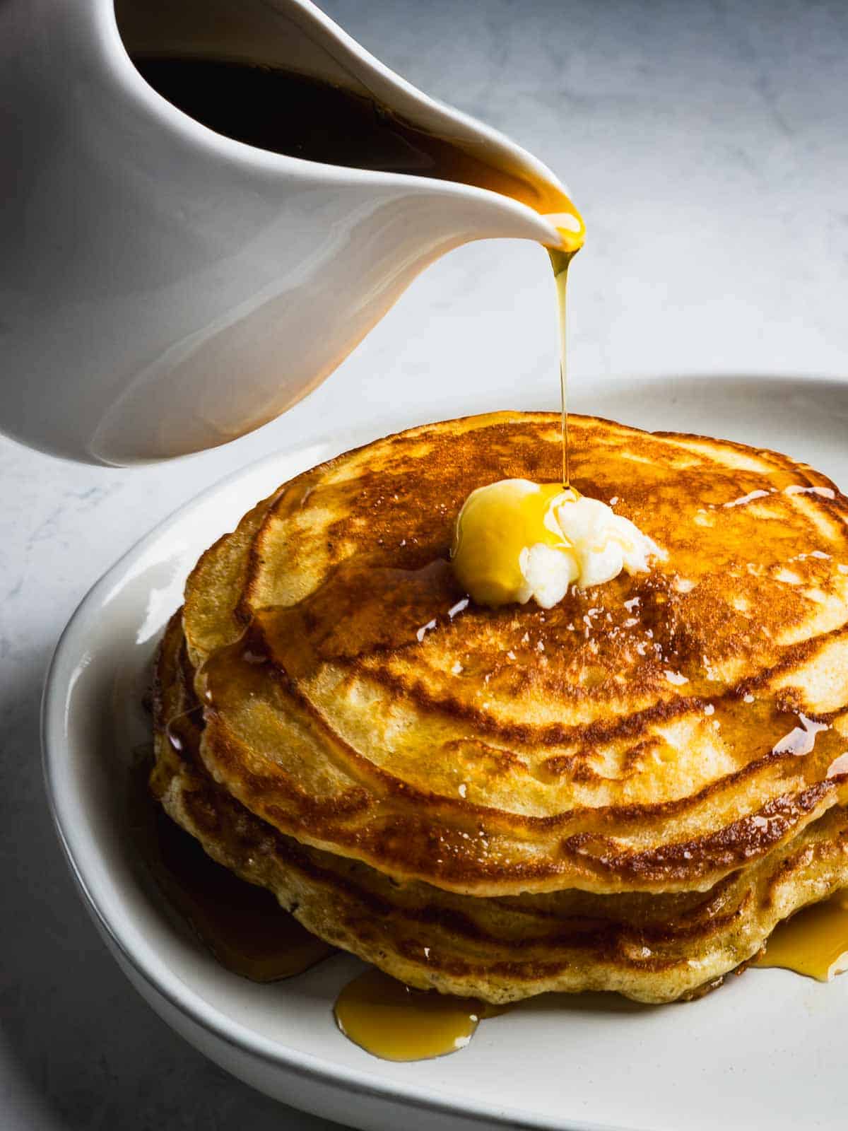 A pitcher pouring warm maple syrup over a stack of crispy buttermilk pancakes with butter on a white plate.