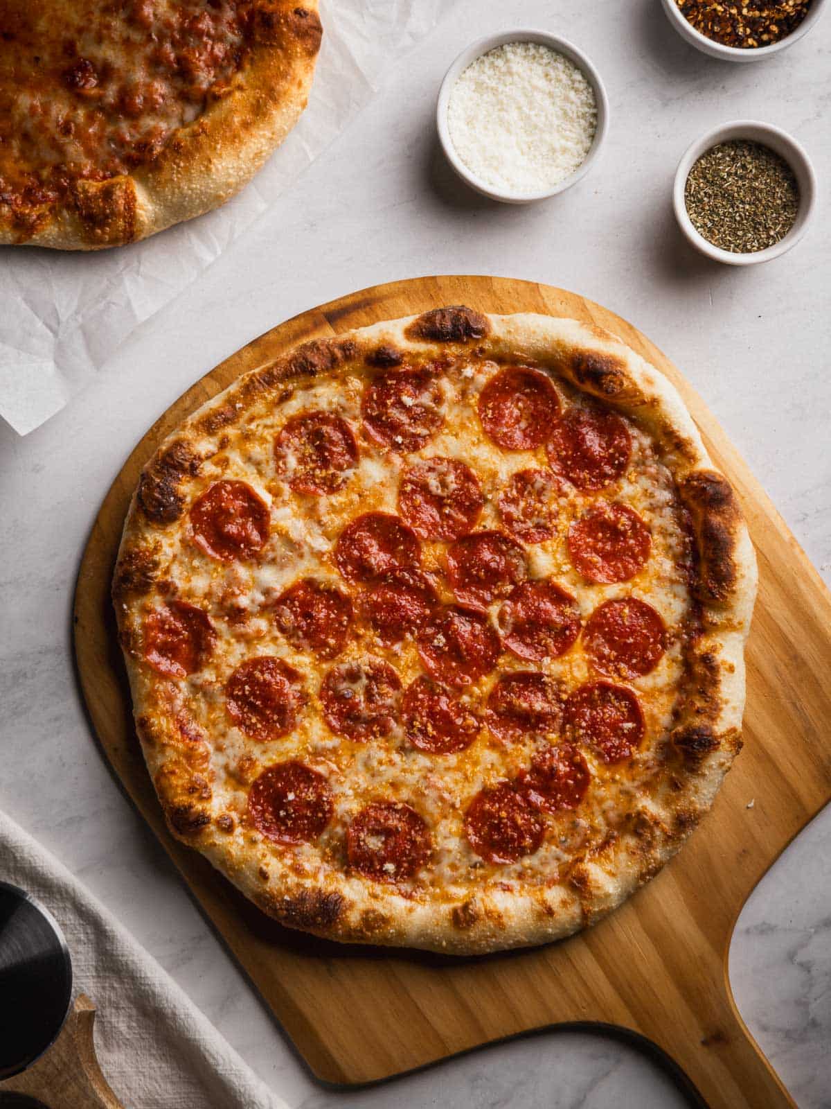 Pepperoni pizza made with cold ferment pizza dough on a wooden pizza peel with cheese, oregano, and red pepper flakes on the side.