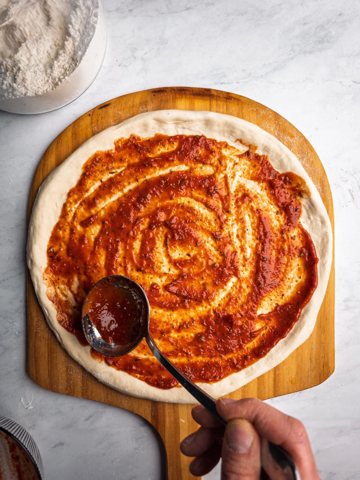 Go-To Homemade Pizza Sauce spread over stretched pizza dough on a wooden peel.