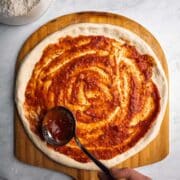 Go-To Homemade Pizza Sauce spread over stretched pizza dough on a wooden peel.
