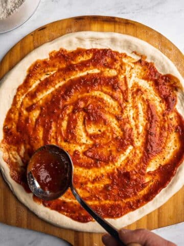 Go-To Homemade Pizza Sauce spread over stretched pizza dough on a wooden peel.