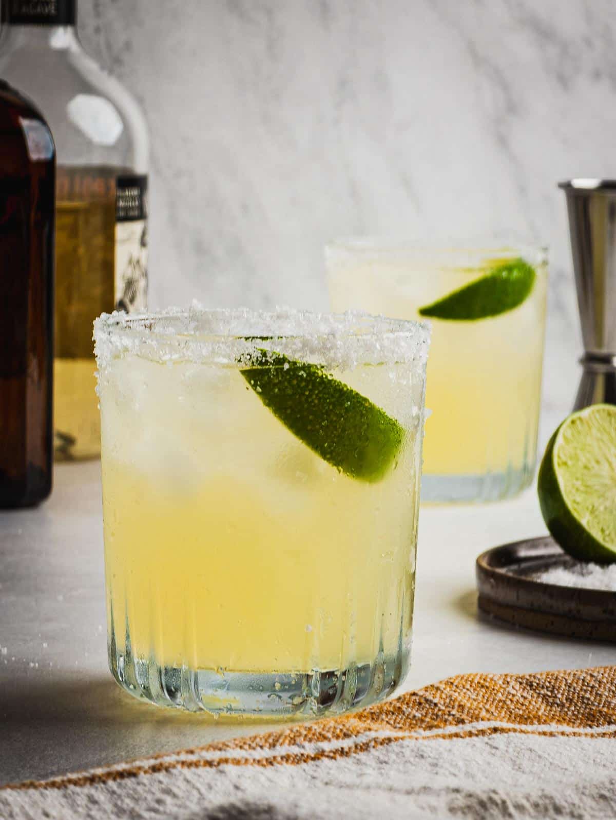 Two glasses with salted rims serving Reposado Margaritas with fresh lime wedges.