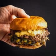 A hand holding a smash burger with onion and jalapeño, topped with pickled okra burger sauce.