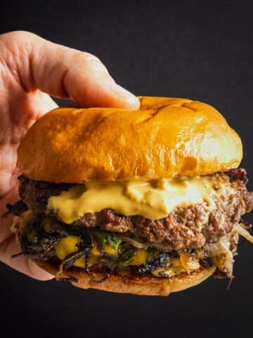 A hand holding a smash burger with onion and jalapeño, topped with pickled okra burger sauce.