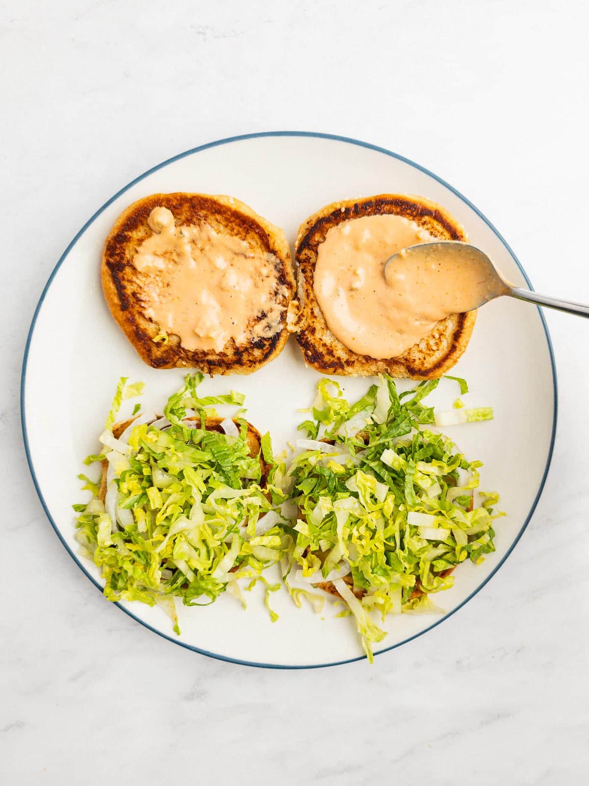 A spoon adding burger sauce to top buns with toppings on the bottom buns.
